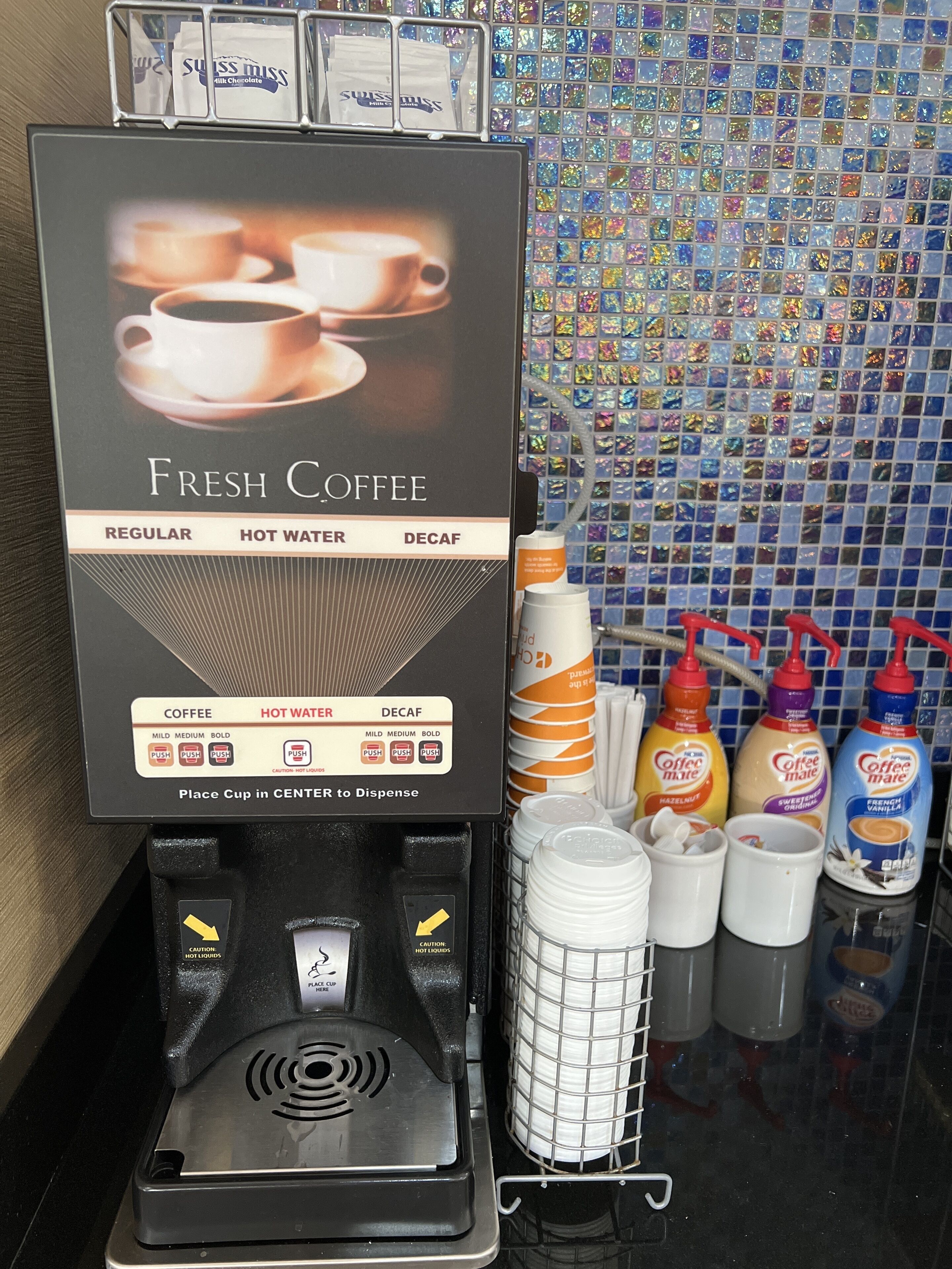 Máquina de café con opciones de café regular, agua caliente y café descafeinado. Tiene un cartel que indica cómo dispensar café y una advertencia sobre líquidos calientes.