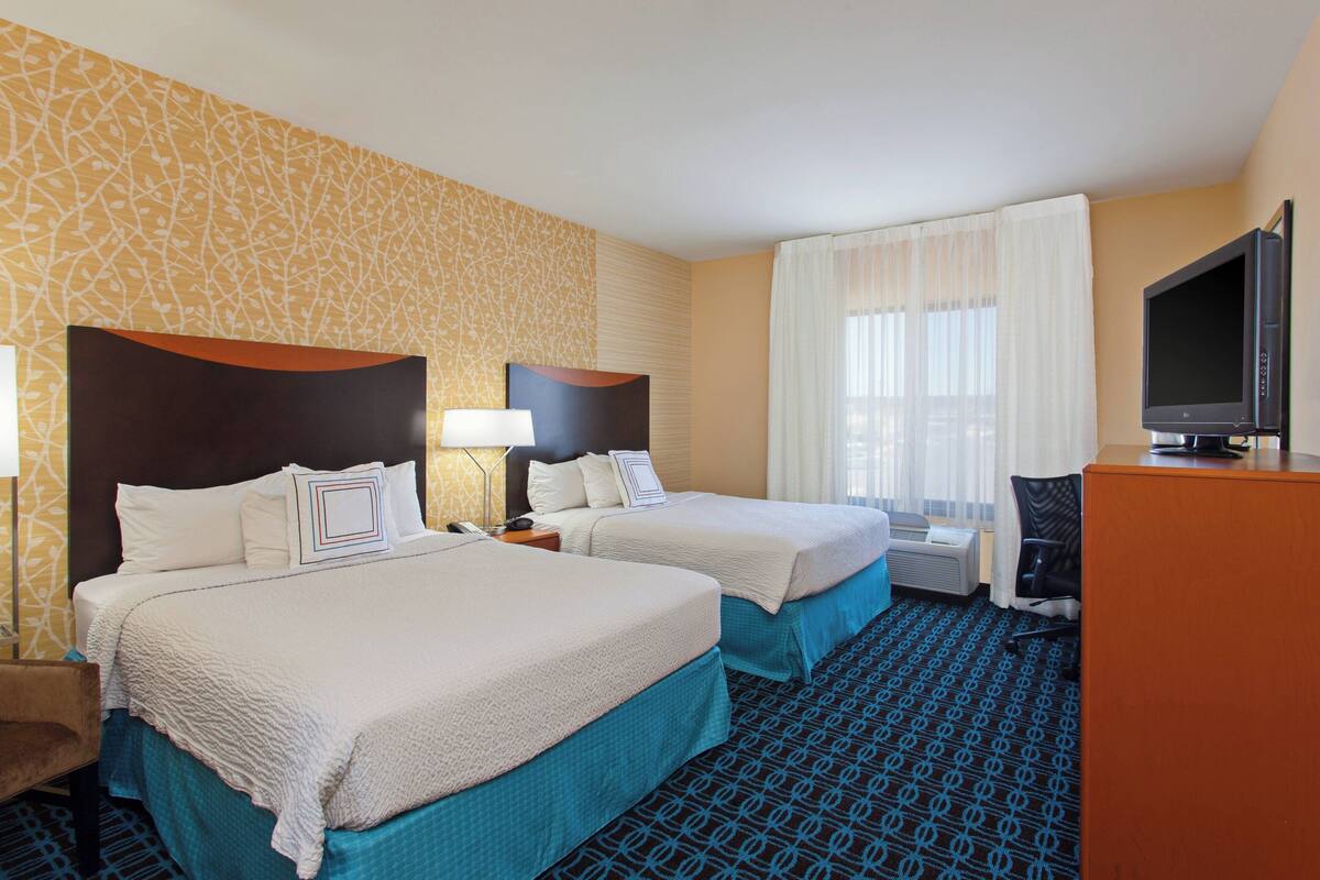 A hotel room with two beds, a desk with a TV, and a window with curtains.