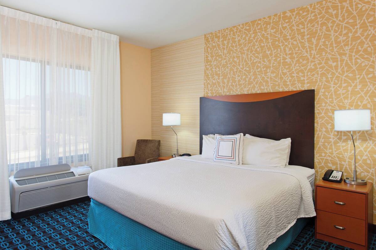 A hotel room with a large bed, a bedside table with a lamp, an air conditioning unit, and a patterned wall.
