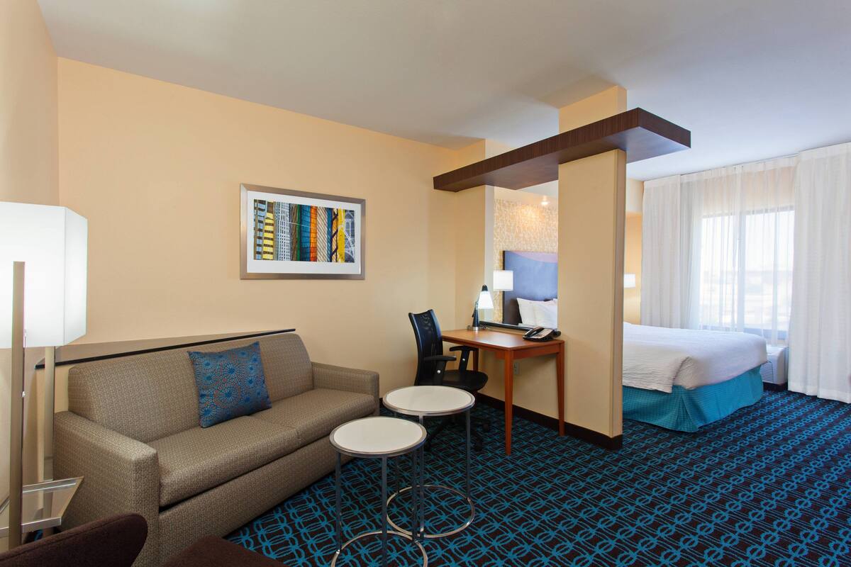 A hotel room with a sofa, two small tables, a desk with a lamp, a bed, and a window with curtains.