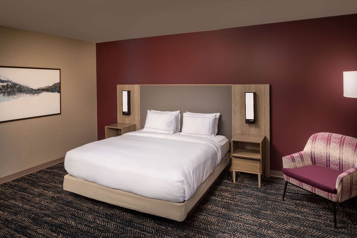 Une chambre d’hôtel avec un grand lit, une chaise, une table de chevet et une lampe fixée au mur.