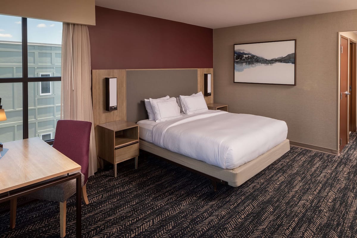 Une chambre d’hôtel avec un grand lit, un bureau, une chaise, une table de chevet, un téléviseur fixé au mur et un tableau encadré accroché au mur.