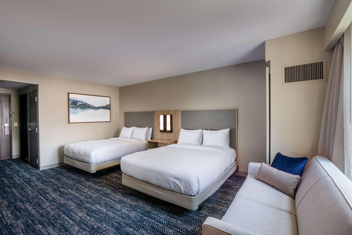 A hotel room with two beds, a sofa, a large window, and a picture on the wall.