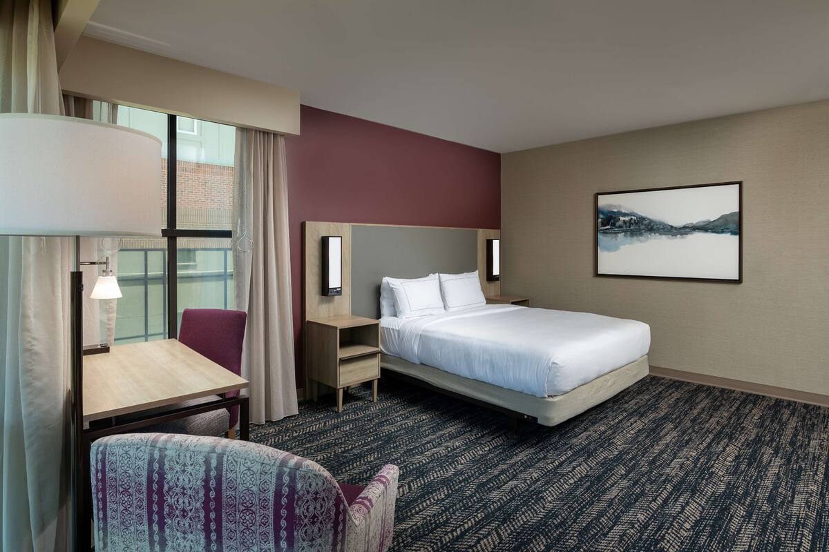 Une chambre d’hôtel avec un lit, un bureau, une chaise, une table de chevet et une fenêtre avec des rideaux.