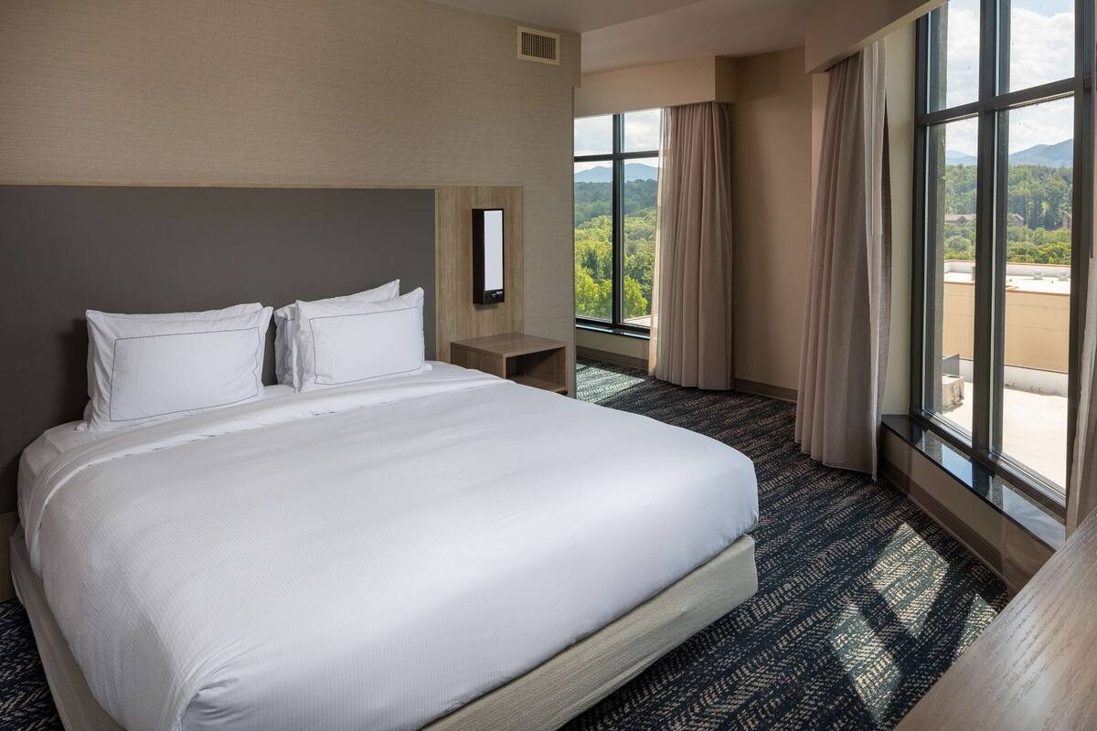 Une chambre d’hôtel avec un grand lit, une table de chevet, un miroir et une fenêtre donnant sur un paysage verdoyant et montagneux.