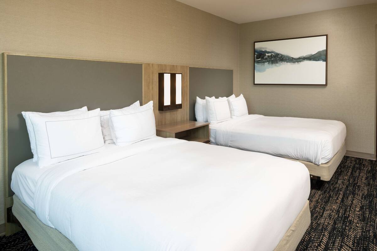 Une chambre d’hôtel avec deux lits, une table de chevet, une lampe fixée au mur et un tableau encadré.