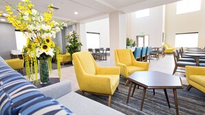 Een hotel lobby met gele fauteuils, een bank en een grote bloemstelling. Er staan tafels en stoelen klaar voor diner of vergaderingen.