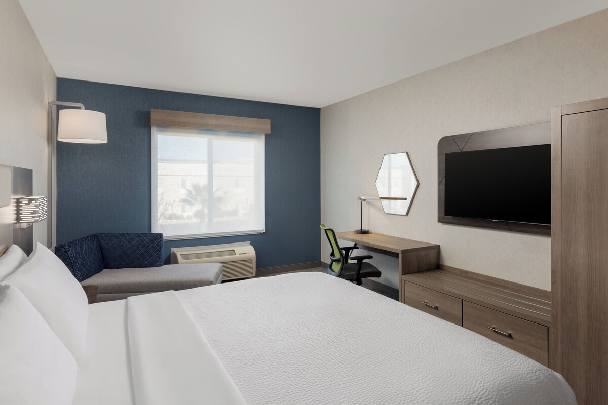 Ein Hotelzimmer mit einem großen Bett, einem Schreibtisch mit Stuhl, einem Flachbildfernseher und einem Fenster mit Jalousien.