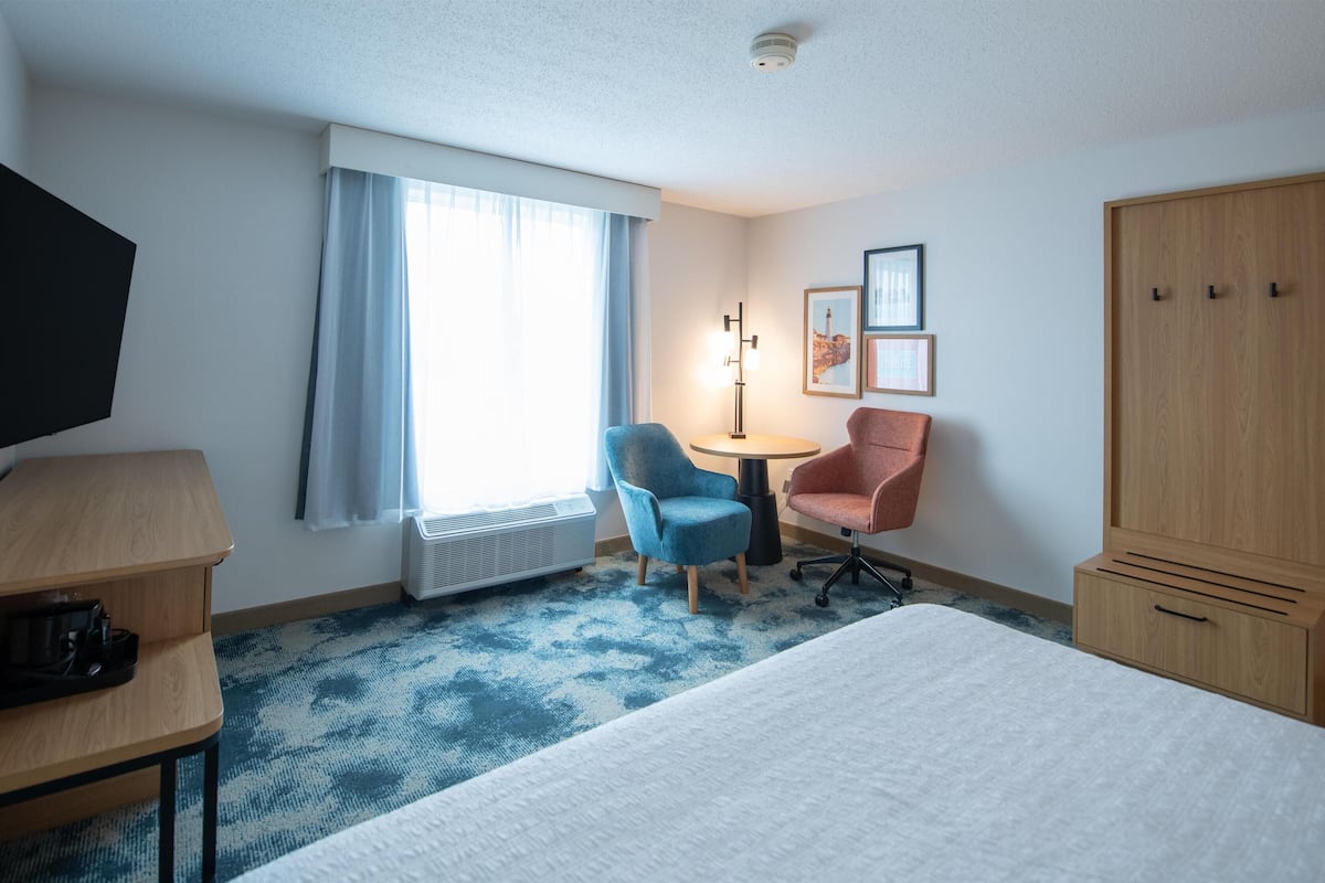 A hotel room with a bed, a desk with a lamp, a chair, a small table, a wardrobe, and a window with curtains.