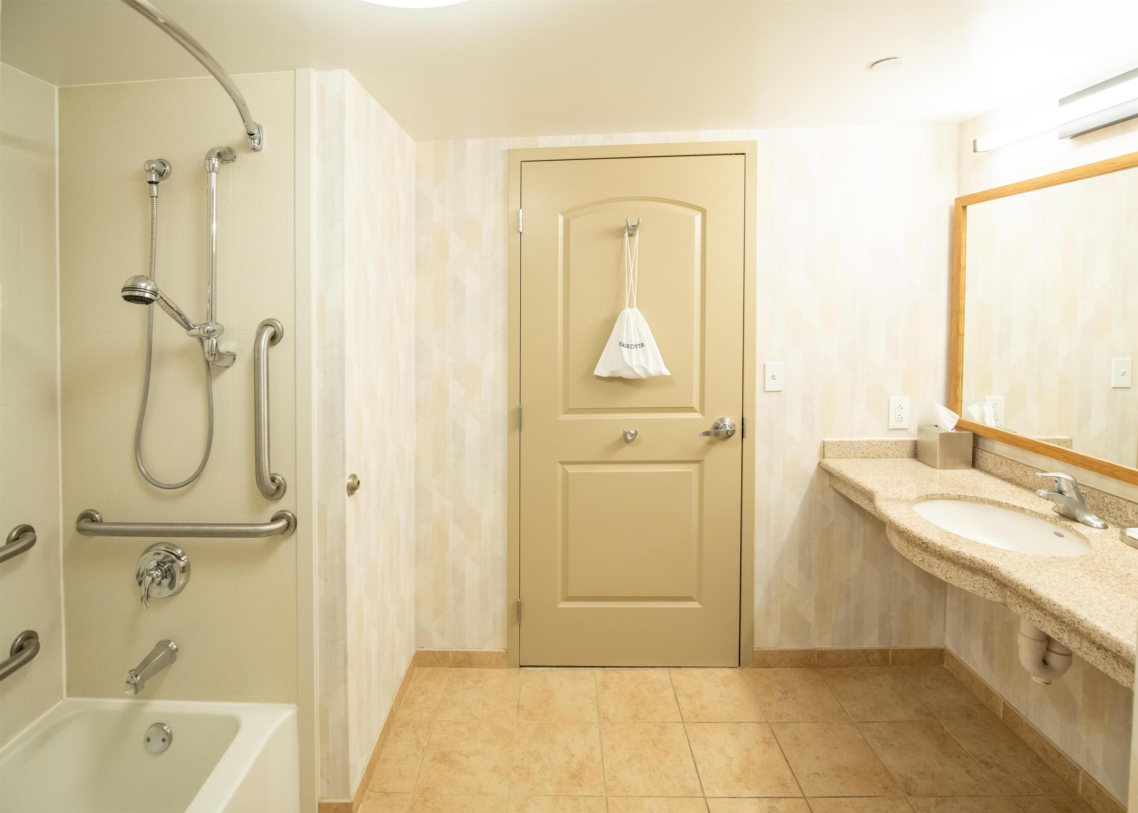 A bathroom with a bathtub, a shower with grab bars, a door with a bag hanging on it, and a sink with a mirror.