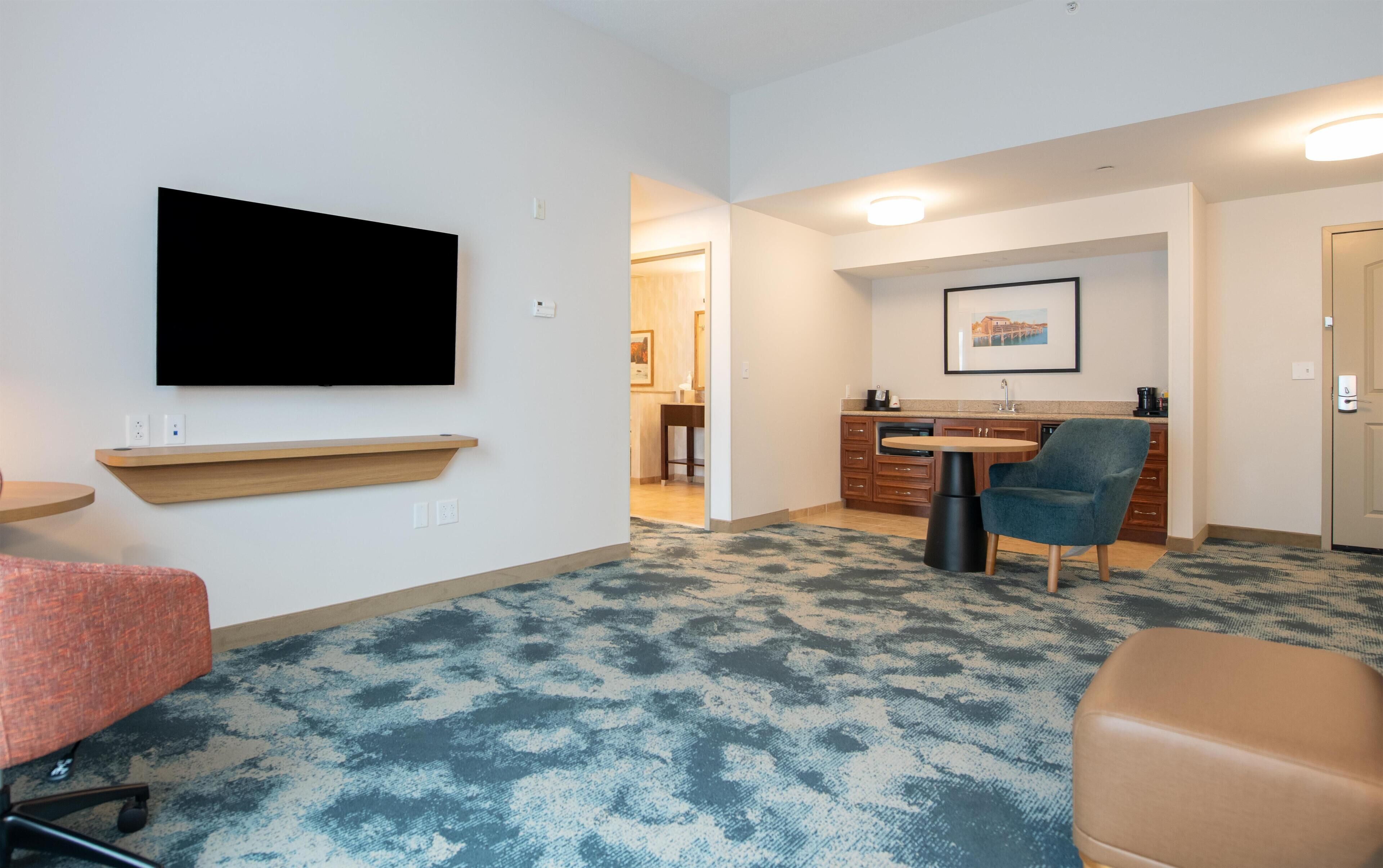 Una habitación de hotel moderna con televisión de pantalla plana, un pequeño estante de madera, una mesa de comedor con sillas, una zona de cocina y una alfombra estampada.