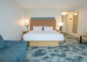 Una habitación de hotel con una cama, mesitas de noche, una lámpara, un espejo y un sillón azul.