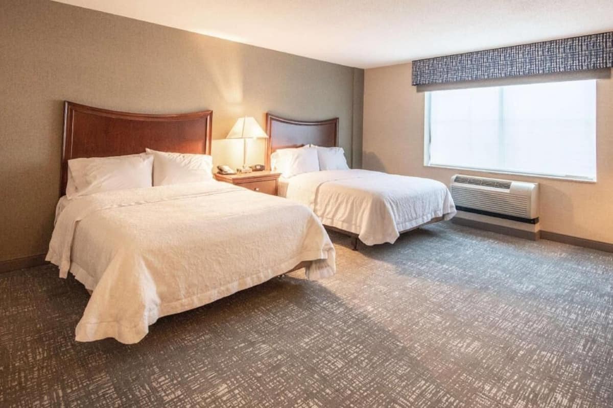 A hotel room with two beds, a nightstand with a lamp, a window with blinds, and a wall-mounted air conditioner.