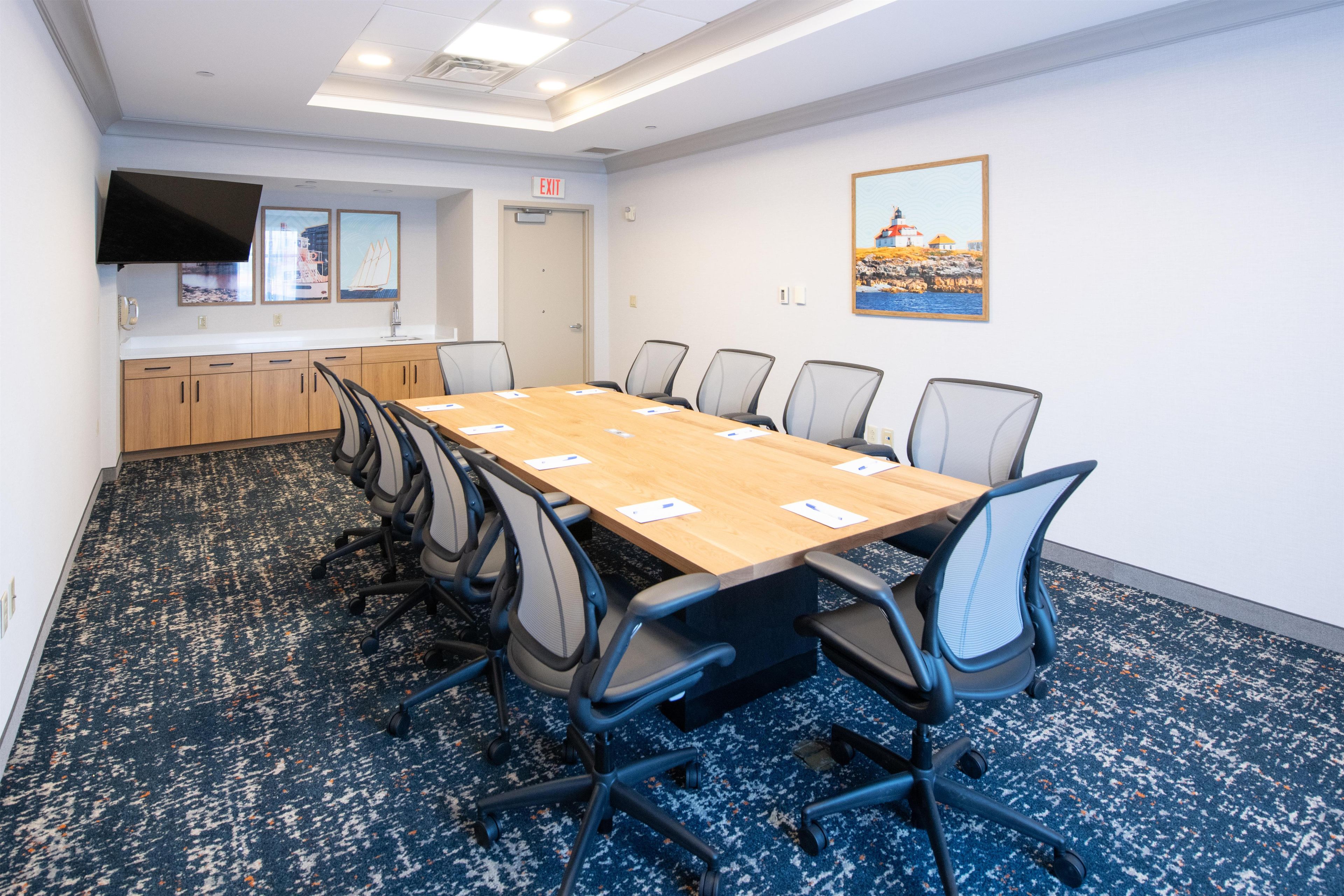 A conference room with a long table, chairs, a kitchen area, and a painting on the wall.