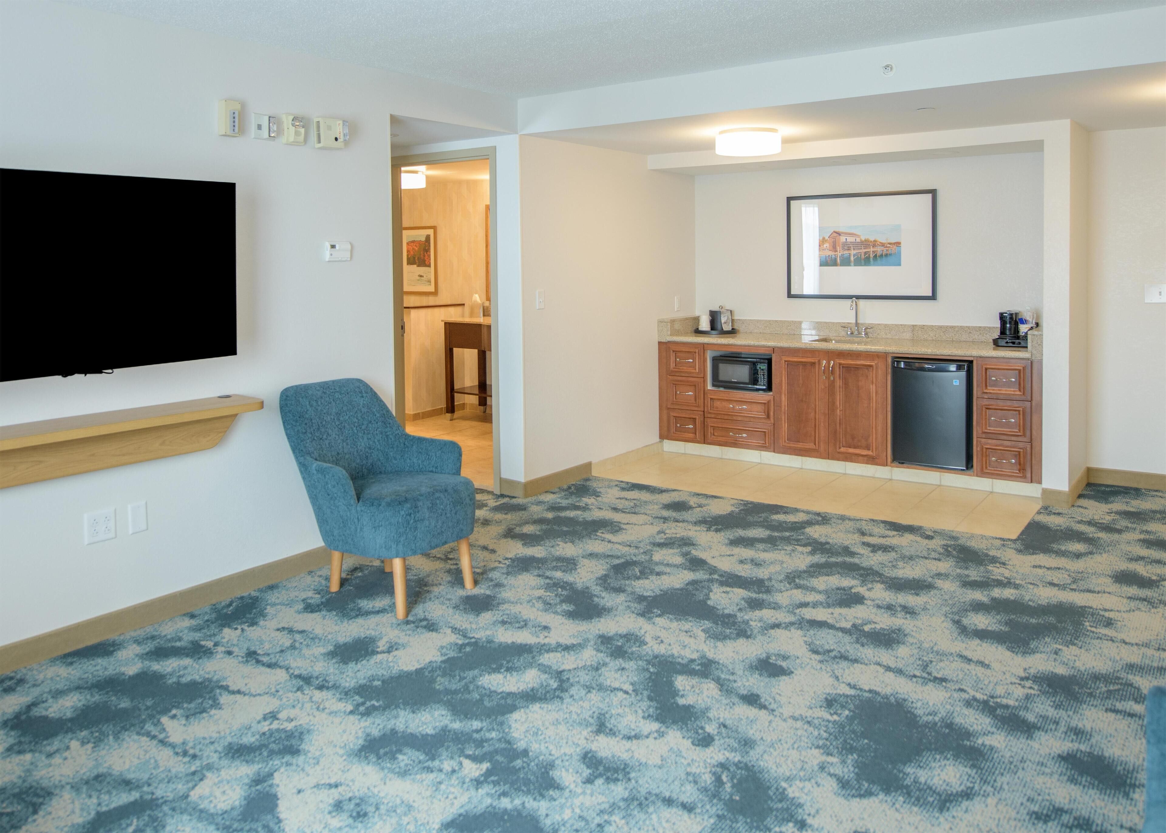 A hotel room with a flat-screen TV mounted on the wall, a blue armchair, a kitchenette with a microwave and dishwasher, and a patterned carpet.