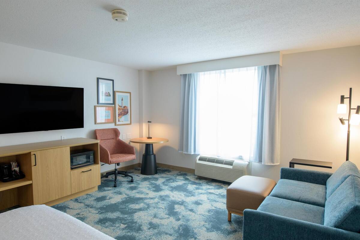 A hotel room with a bed, a desk, a chair, a sofa, a TV, a window with curtains, and a floor lamp.