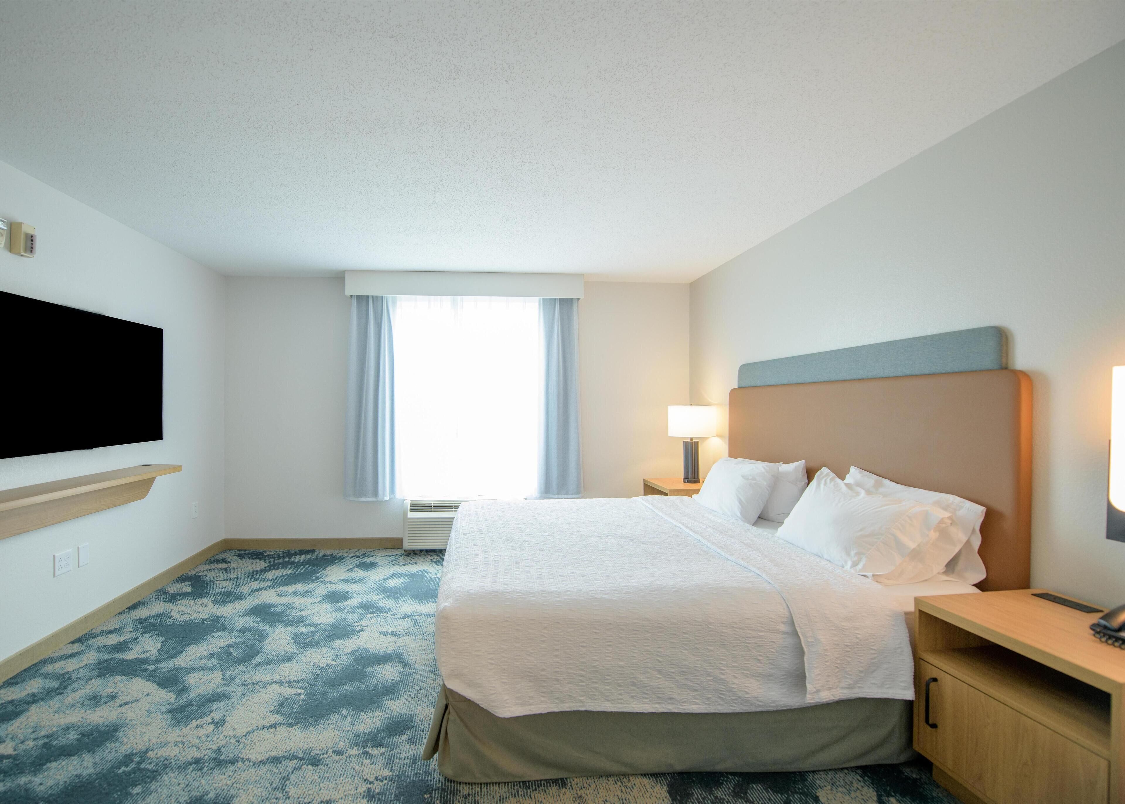 A hotel room with a bed, a TV mounted on the wall, a window with curtains, a bedside table with a lamp, and a carpeted floor.
