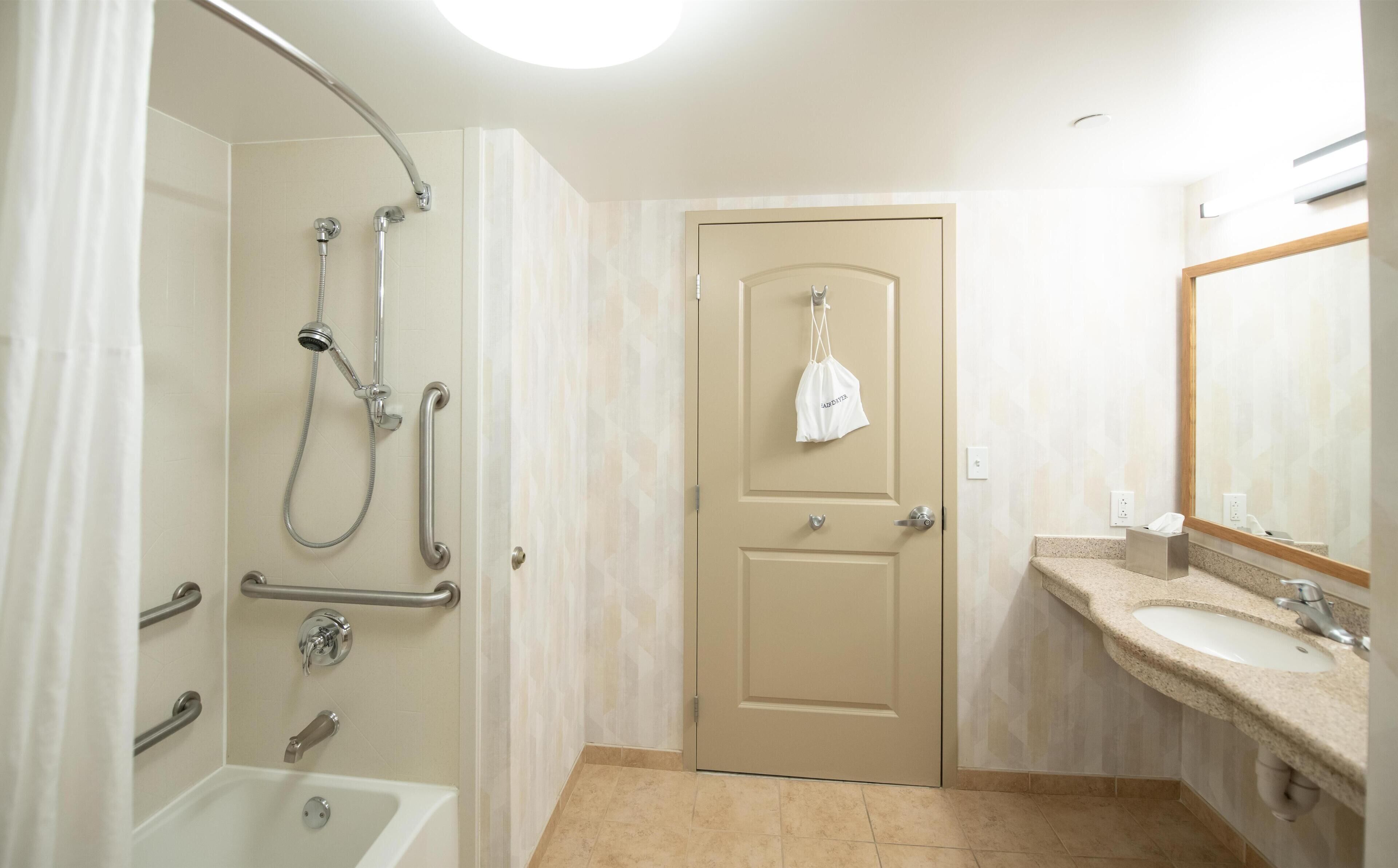 Een badkamer met een douche, handgrepen, een deur met een handdoek eraan, een wastafel en een spiegel.