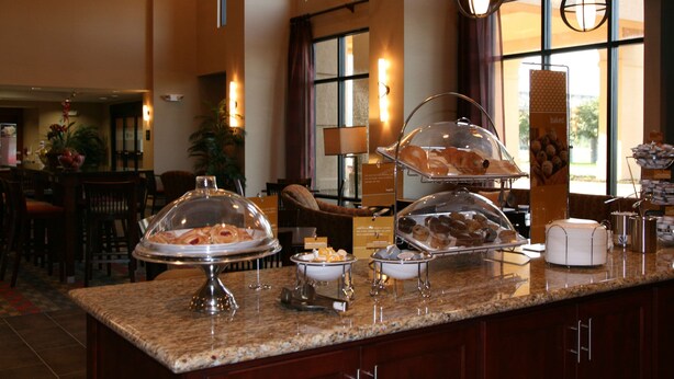 A buffet counter with various food items under glass domes, a coffee station, and a seating area with chairs and a table. - Hampton Inn & Suites West Point (West Point)