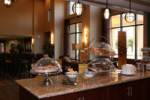 A buffet counter with various food items under glass domes, a coffee station, and a seating area with chairs and a table. - Hampton Inn & Suites West Point (West Point)