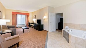 A hotel room with a living area, a TV, a desk, a sofa, a bathroom with a bathtub, and a door leading to another room. - La Quinta Inn & Suites by Wyndham Stillwater-University Area (Stillwater)