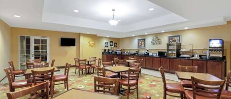 A hotel dining area with multiple tables and chairs, a kitchen counter, and a television.