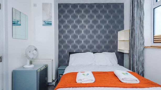 Une chambre d’hôtel comprenant un lit, une table de chevet, un radiateur, un ventilateur fixé au mur et une fenêtre avec des rideaux.