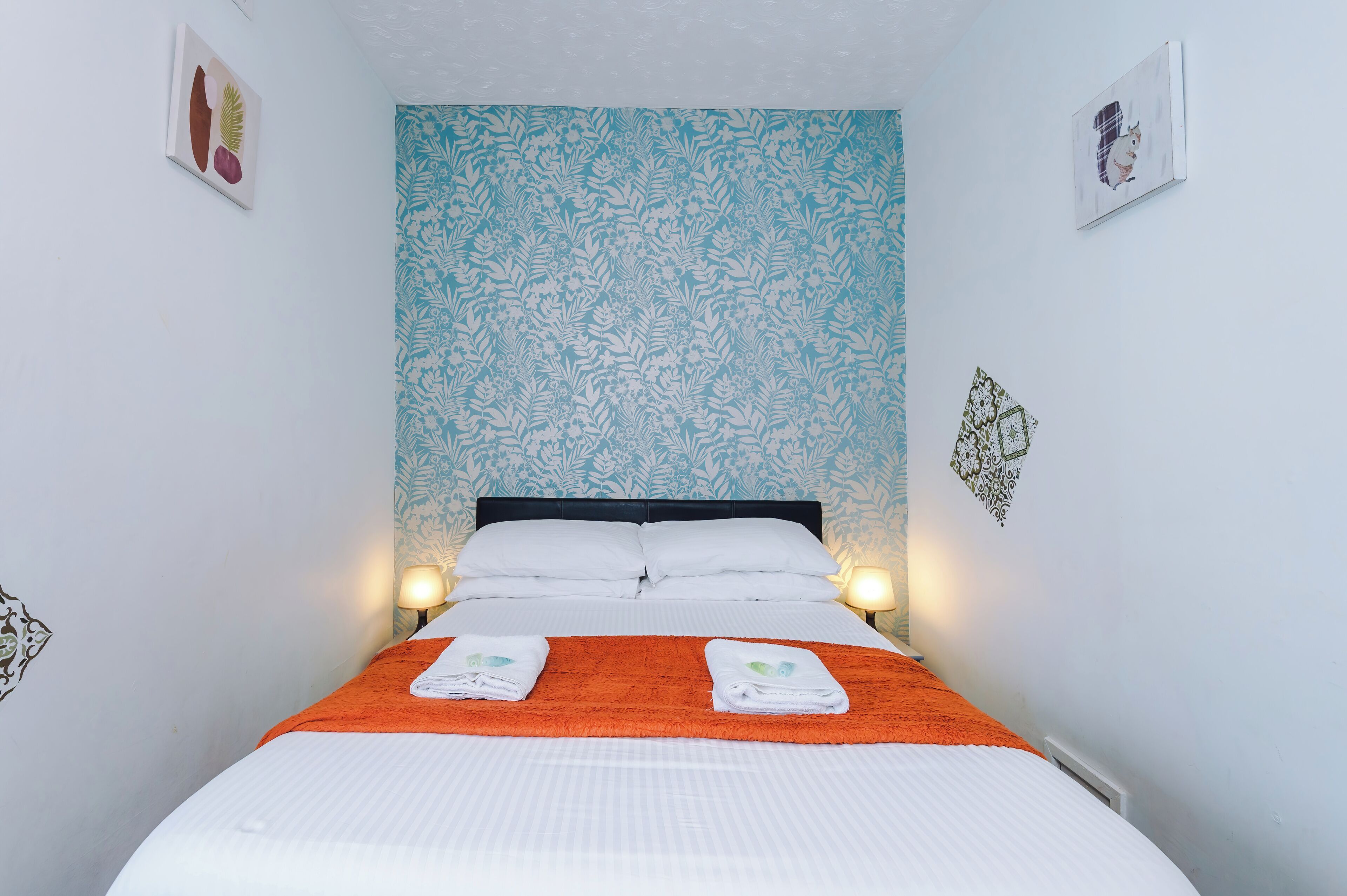 A neatly made bed with white linens and an orange throw blanket, a black headboard, and a floral-patterned wall.