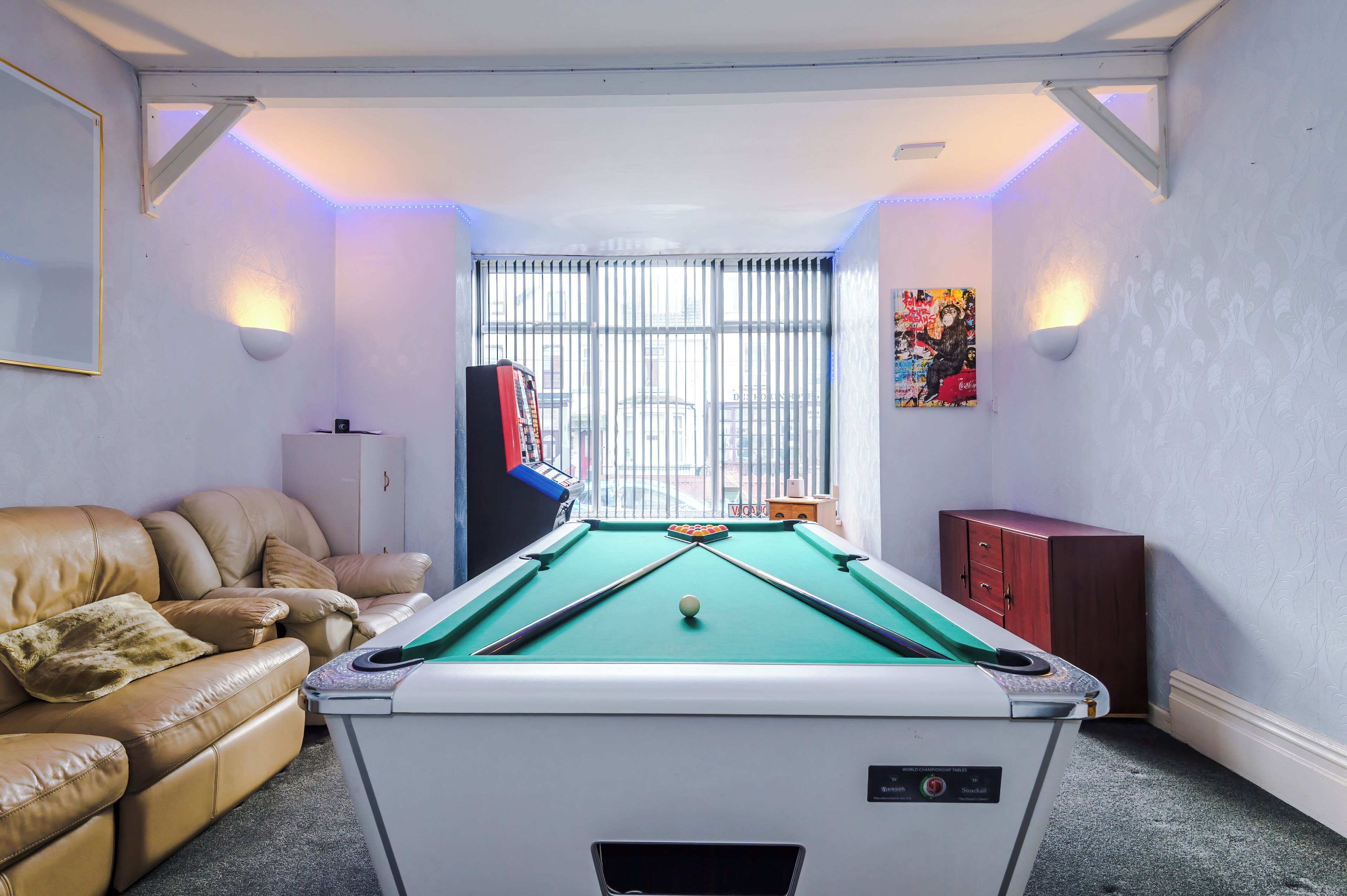 A pool table with a green felt surface, a leather sofa, and a window with vertical blinds.