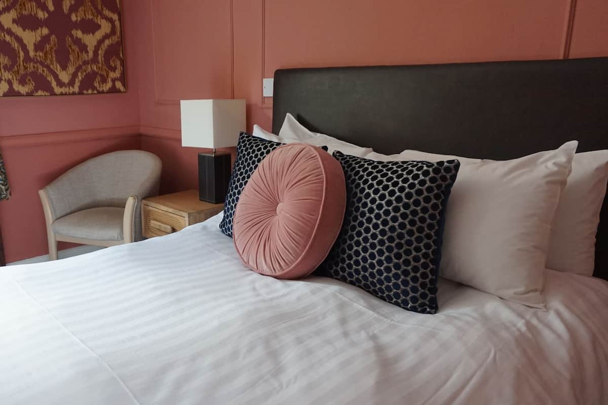 A neatly made bed with white linens, a headboard, and decorative pillows. There is a chair and a nightstand with a lamp in the room.