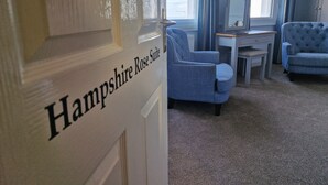 A room with a sign reading "Hampshire Rose Suite," a blue armchair, a small table with a lamp, and a window with curtains. - Dunkerleys Hotel (Deal)