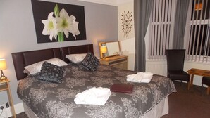 A hotel room with a large bed, a bedside table with a lamp, a chair, a window with curtains, and a wall with a floral painting. - Easton Court (Paignton)