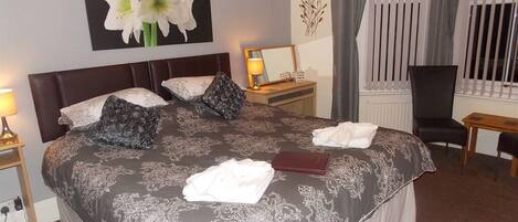 A hotel room with a large bed, a bedside table with a lamp, a chair, a window with curtains, and a wall with a floral painting.