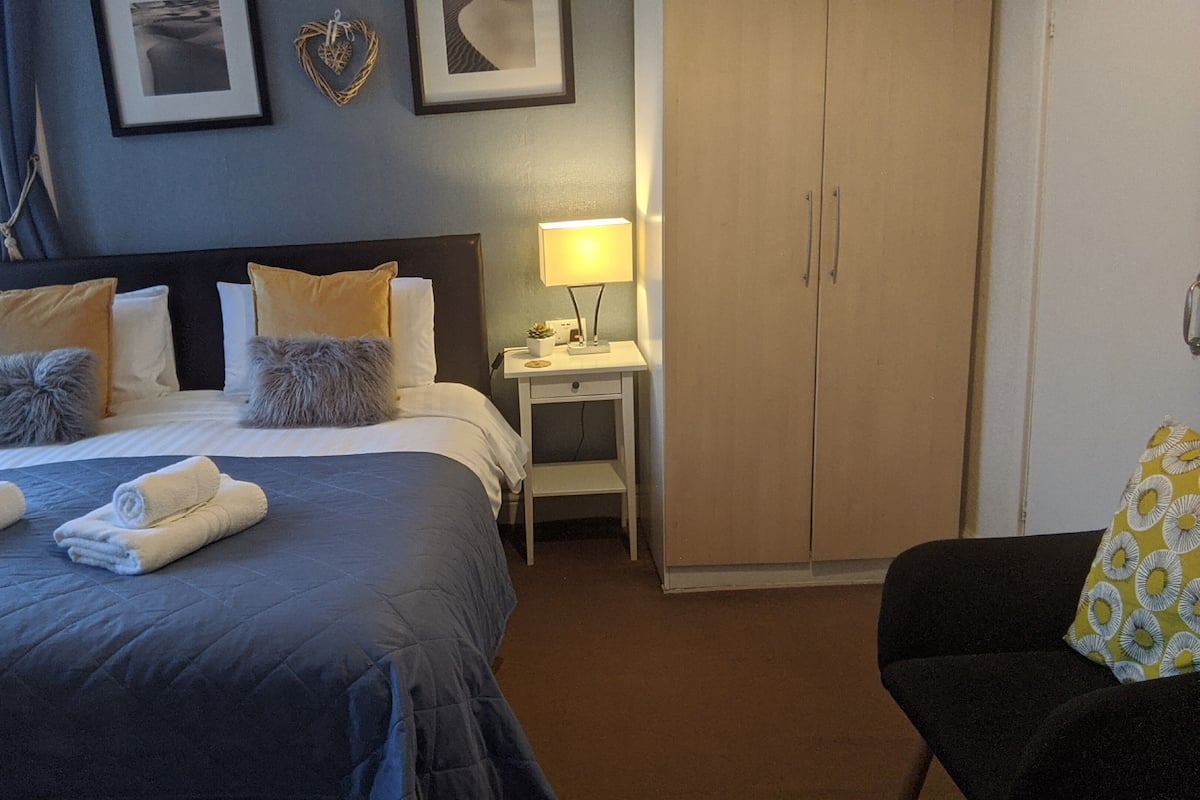 Cama bem arrumada com toalhas, mesa de cabeceira com abajur, guarda-roupa e uma cadeira.