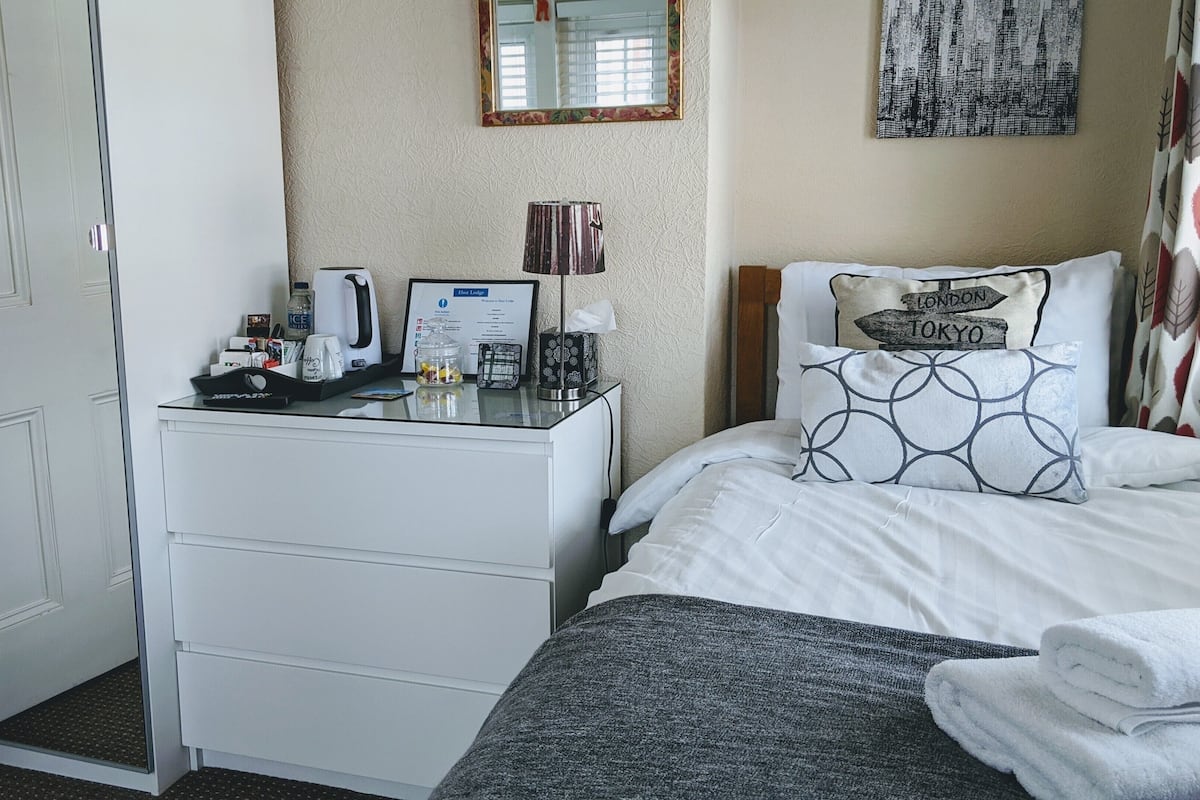 Quarto de hotel com cama, escrivaninha com cafeteira, espelho e placa de boas-vindas emoldurada.