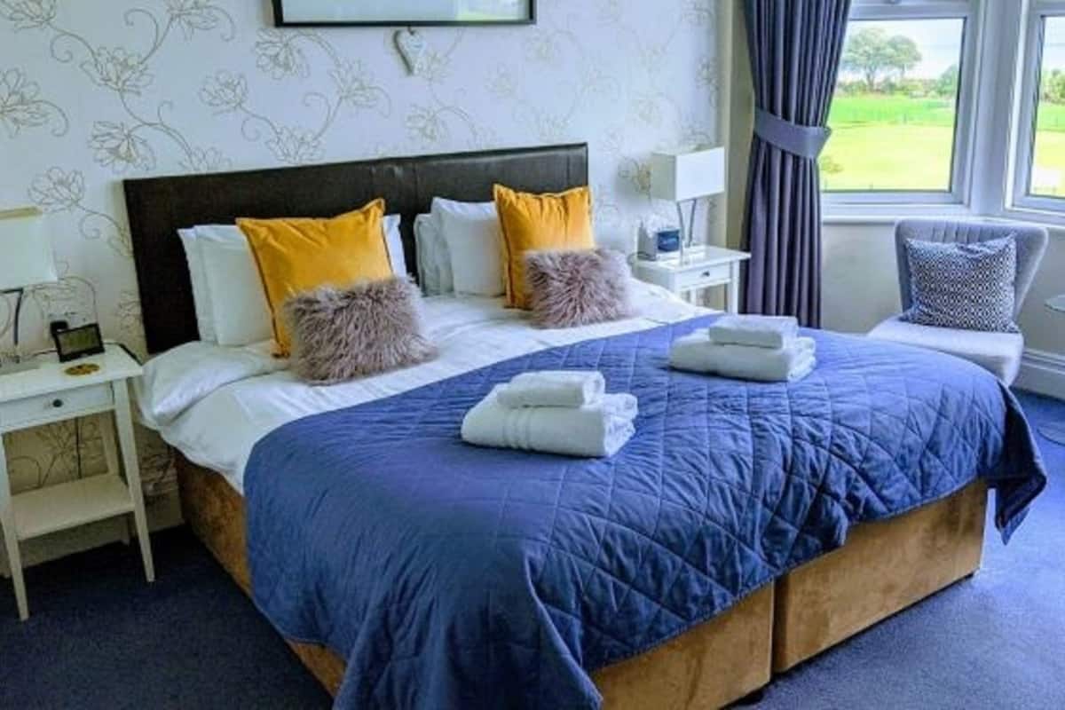 Cama bem arrumada com colcha azul, duas almofadas e toalhas dobradas. Tem uma mesa de cabeceira com abajur e uma pequena planta. Uma janela com cortinas mostra uma paisagem verde lá fora.