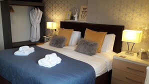 A hotel room with a large bed, two bedside tables, a nightstand, a robe hanging on the wall, and a patterned wallpaper. - Ebor Lodge (Eastbourne)