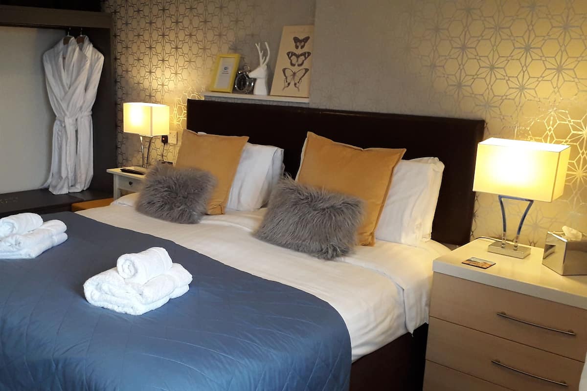 Quarto de hotel com uma cama grande, duas mesas de cabeceira, um criado-mudo, um roupão pendurado na parede e papel de parede estampado.
