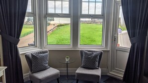 A room with two grey armchairs, a small round table with a plant, and a view of a green lawn through large windows. - Ebor Lodge (Eastbourne)