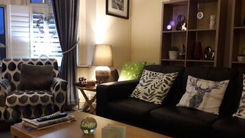 A living room with a patterned armchair, a black sofa with leaf-patterned pillows, a small table with a lamp, and a shelf with decorative items.