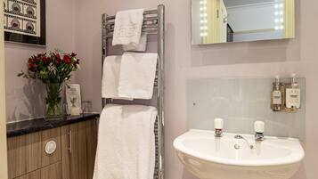 A bathroom with a white toilet, a sink with two faucets, a towel rack with two white towels, a mirror with lights, and a framed picture on the wall.