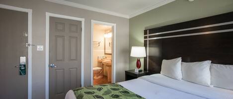 A hotel room with a bed, a nightstand with a lamp, a door leading to a bathroom, and a wall-mounted light fixture.