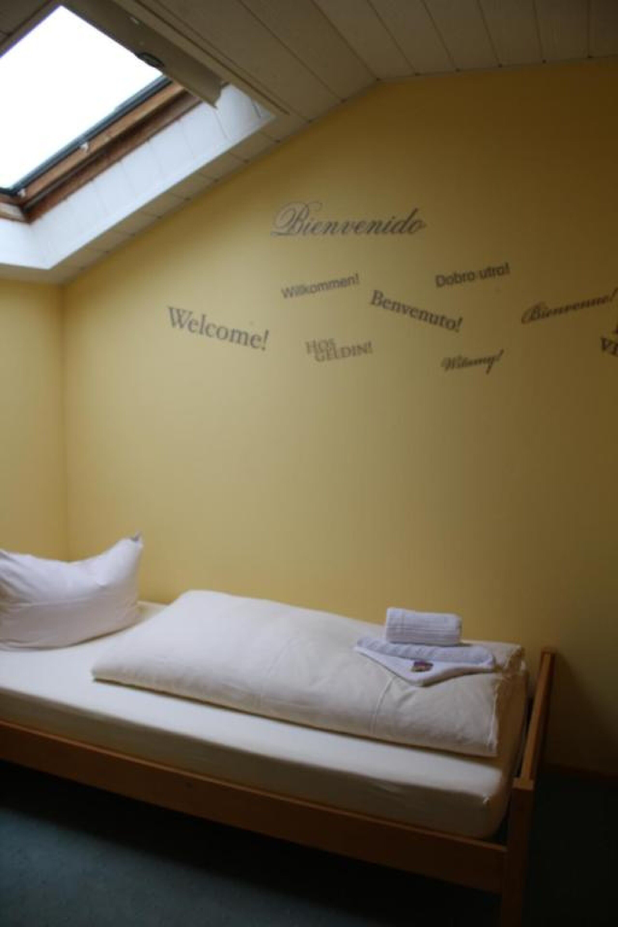 A single bed with a white mattress and pillow, a skylight above, and a wall with 'Welcome!' and 'Bienvenue!' written in multiple languages.