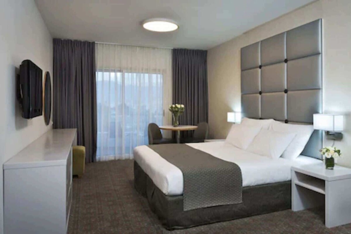 Una habitación de hotel con cama, mesita de noche, lámpara, silla, escritorio y televisor.
