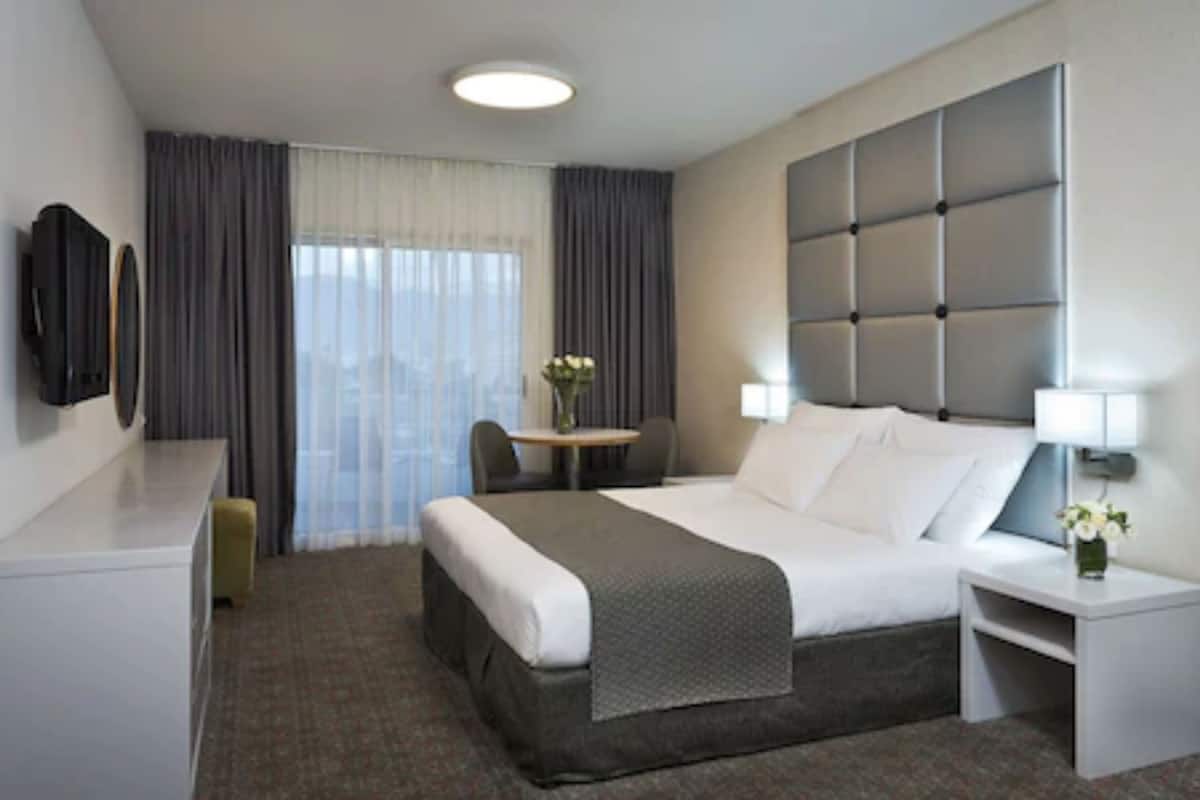 A hotel room with a bed, bedside table, chair, desk, and a TV on the wall.