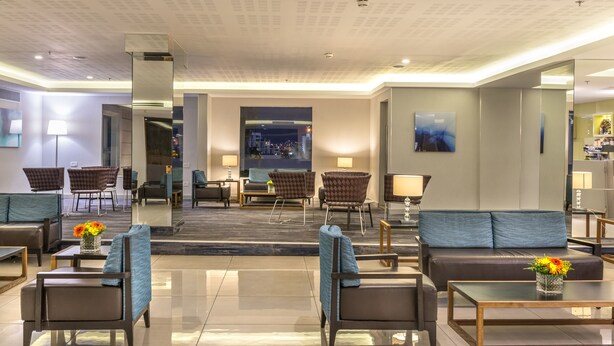 Um saguão de hotel moderno com área de estar, balcão de recepção e vista para a paisagem urbana.