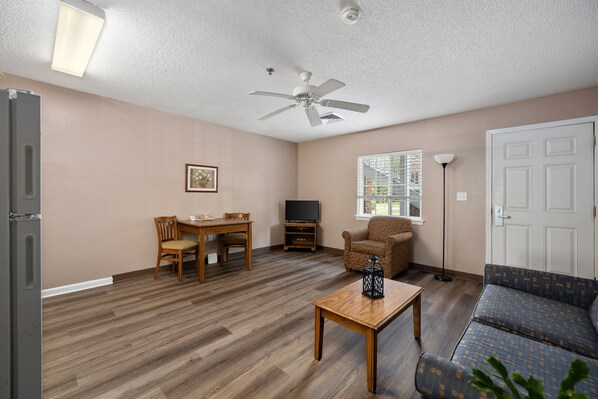 A living room with a ceiling fan, a television, a dining table with chairs, a sofa, and a window with blinds. - Affordable Suites Greenville (Greenville)