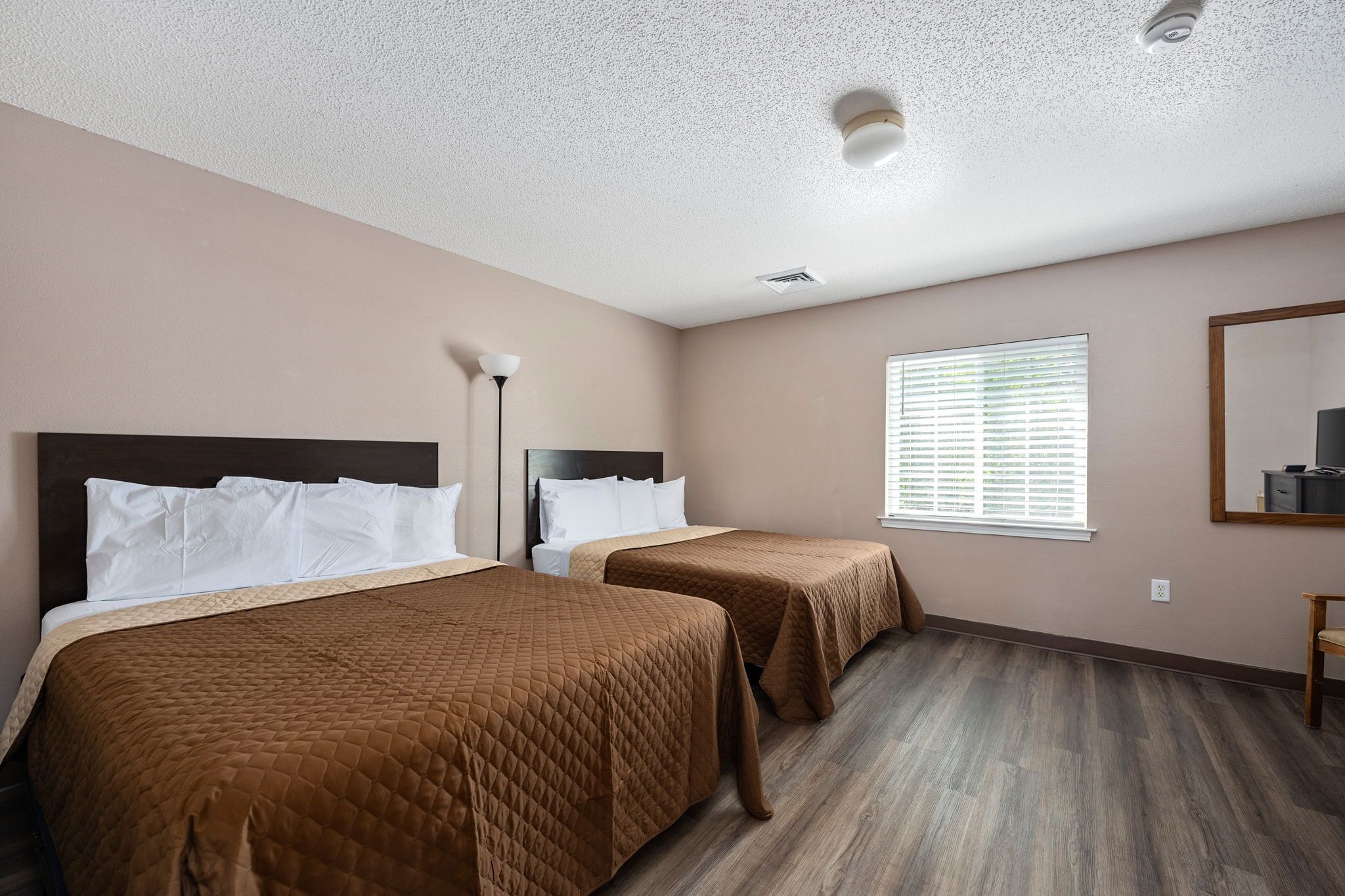 Une chambre d’hôtel avec deux lits, des murs de couleur beige, un plancher en bois, une fenêtre avec des stores et un miroir sur le mur.