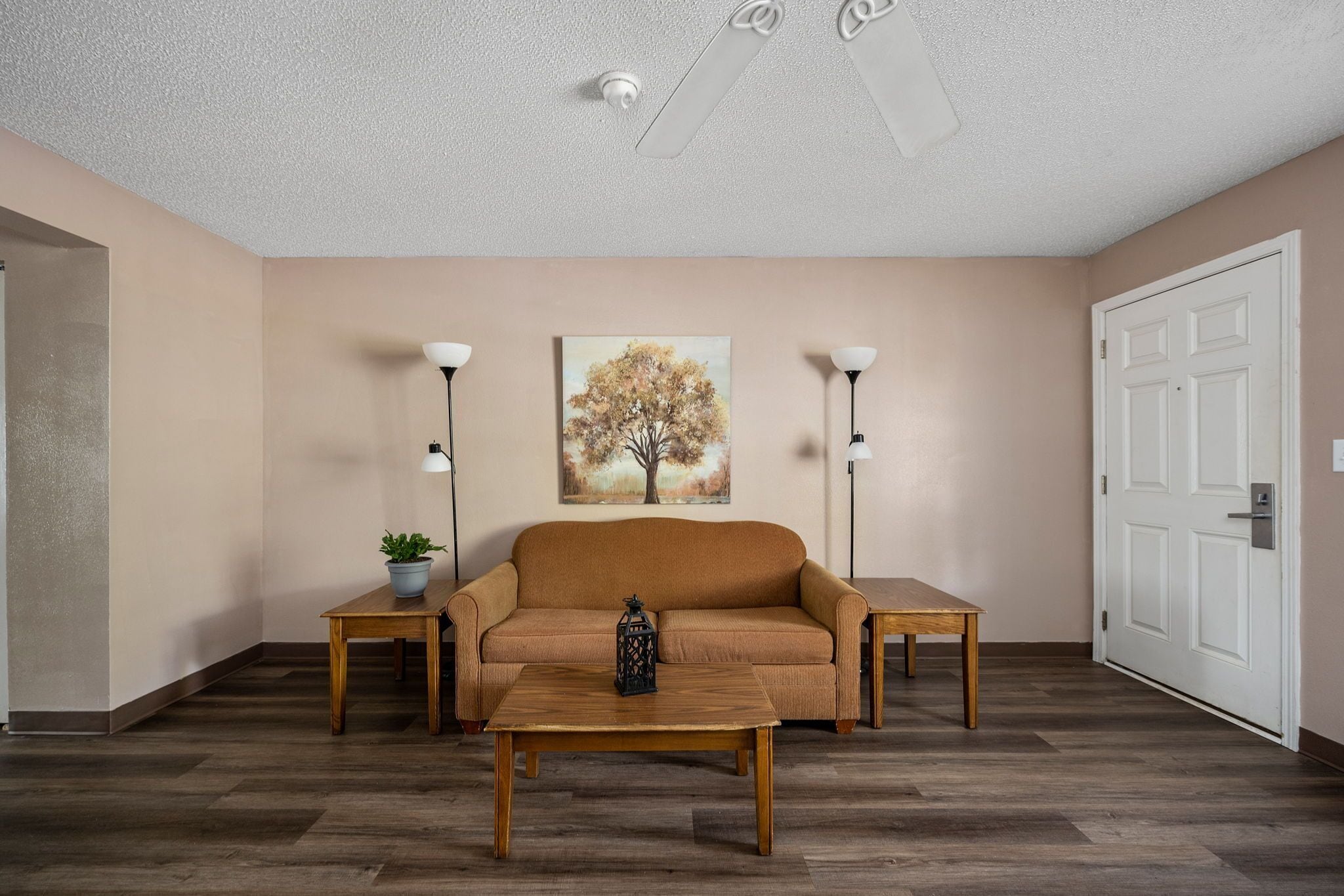 Uma sala de estar com sofá, duas mesas laterais, uma mesa de centro, um quadro e uma porta.