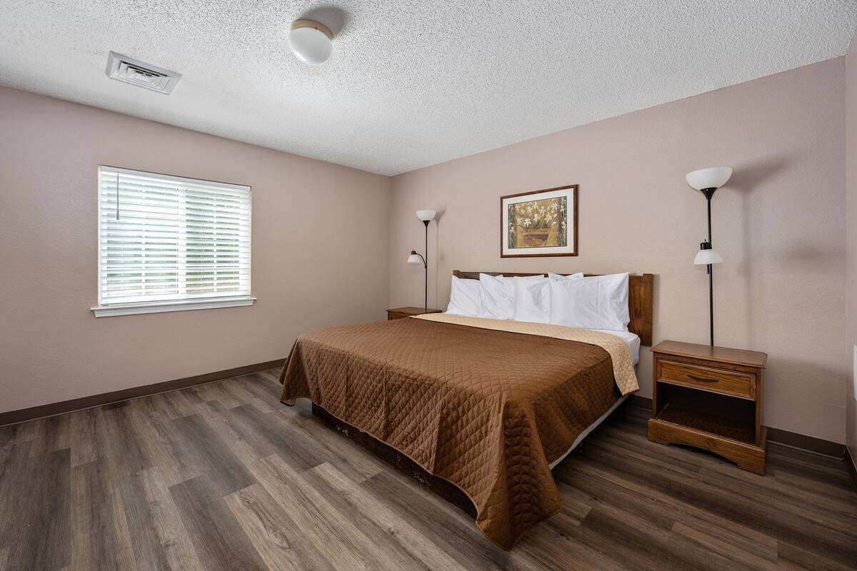A bedroom with a bed, bedside tables, a window with blinds, a ceiling vent, and a wall-mounted picture.